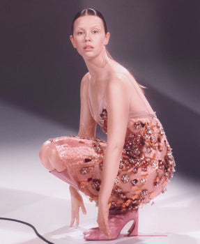 Mia Goth wearing a sheer outfit with floral embellishments on a plain background