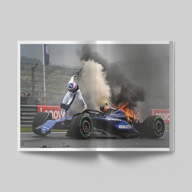 A square art print or photo print showing a dramatic motorsport scene: a blue Formula 1 car on a racetrack with smoke, fire, and a driver in white racing suit leaning on it. The print is presented against a light gray background with a soft shadow.