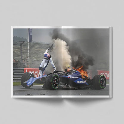 A square art print or photo print showing a dramatic motorsport scene: a blue Formula 1 car on a racetrack with smoke, fire, and a driver in white racing suit leaning on it. The print is presented against a light gray background with a soft shadow.