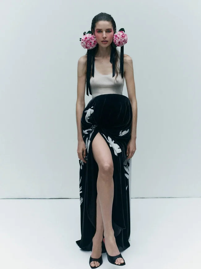 A model stands against a plain white studio background wearing a sleeveless beige top and a long black skirt with white floral print and a high slit.