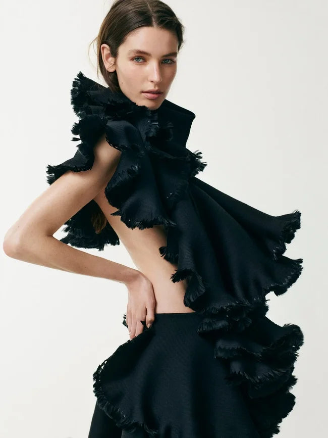 A model wearing a black ruffled dress with a high ruffled collar, a sleeveless back, and a flared skirt, photographed against a plain light background.